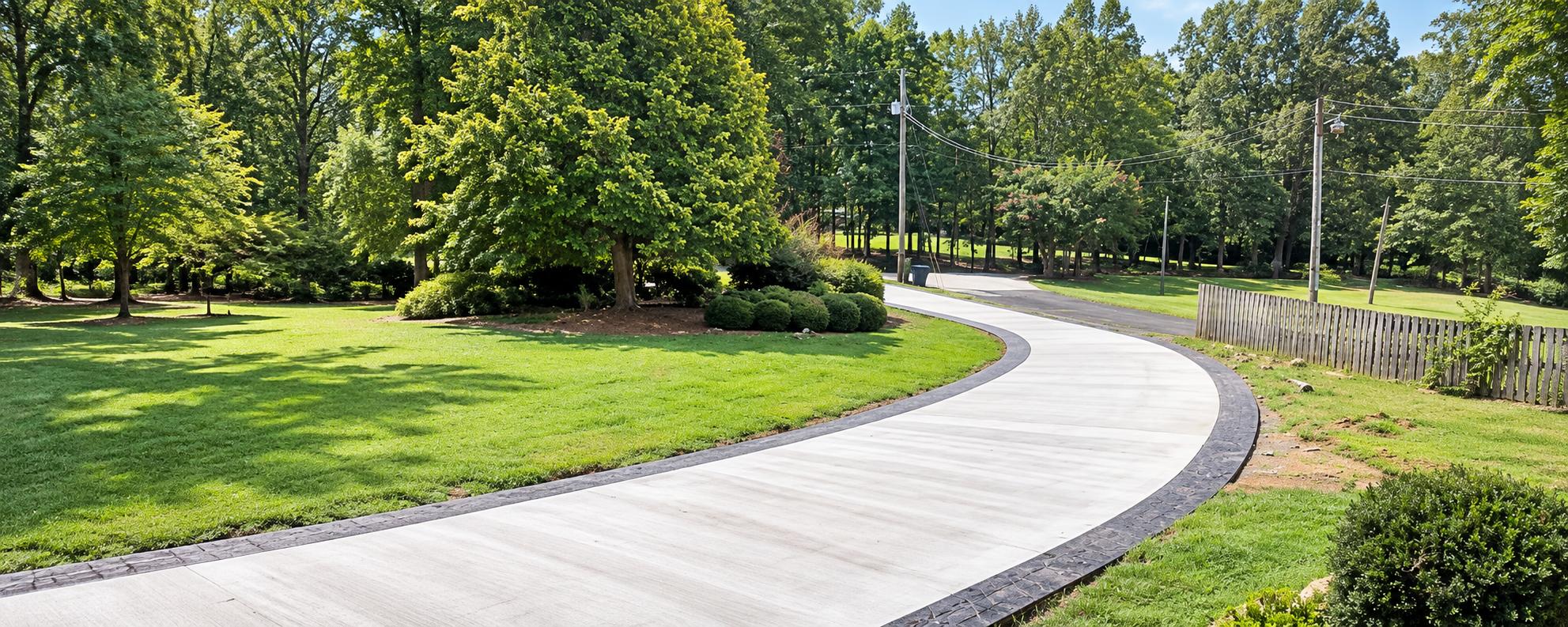 Curved concrete driveway with dark stamped edging beside a wooded residential lawn