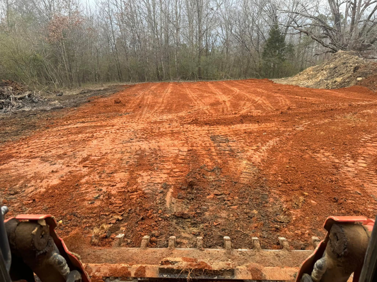 Graded red-clay building pad bordered by trees, viewed from grading equipment